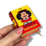 A hand with pink manicured nails holds a miniature hardcover book designed to look like a Peanuts book, featuring the character Mafalda on a yellow and red cover with the Italian text 'Il meglio di MAFALDA'.